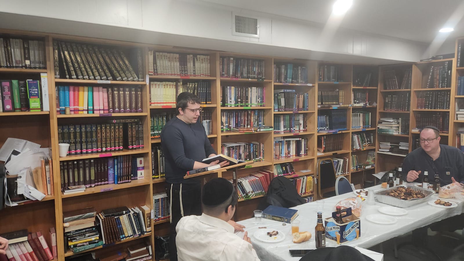 A talmid reading from a sefer at a shtender among the bookshelves