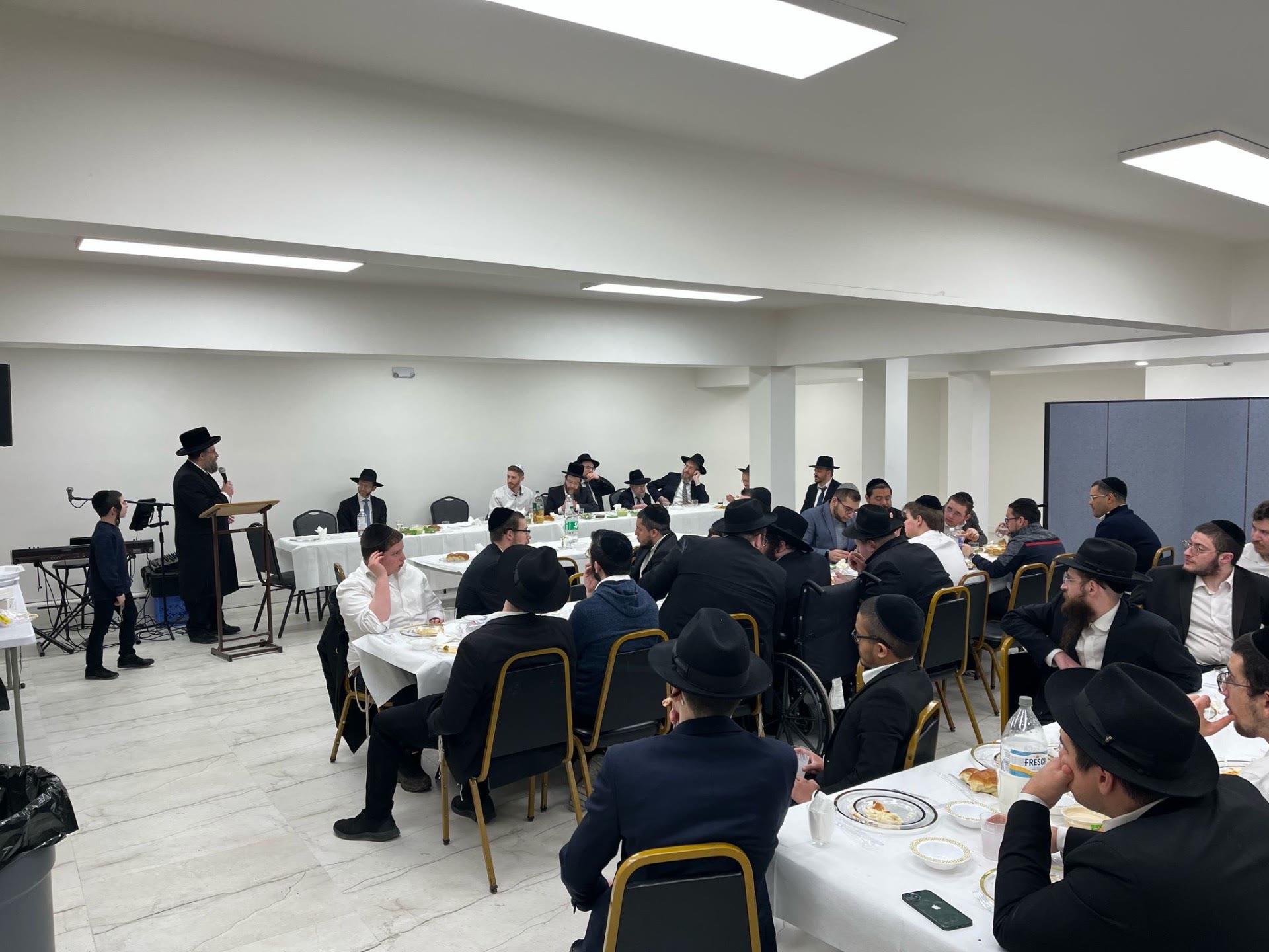 A rebbe speaking at a podium to talmidim seated at a meal