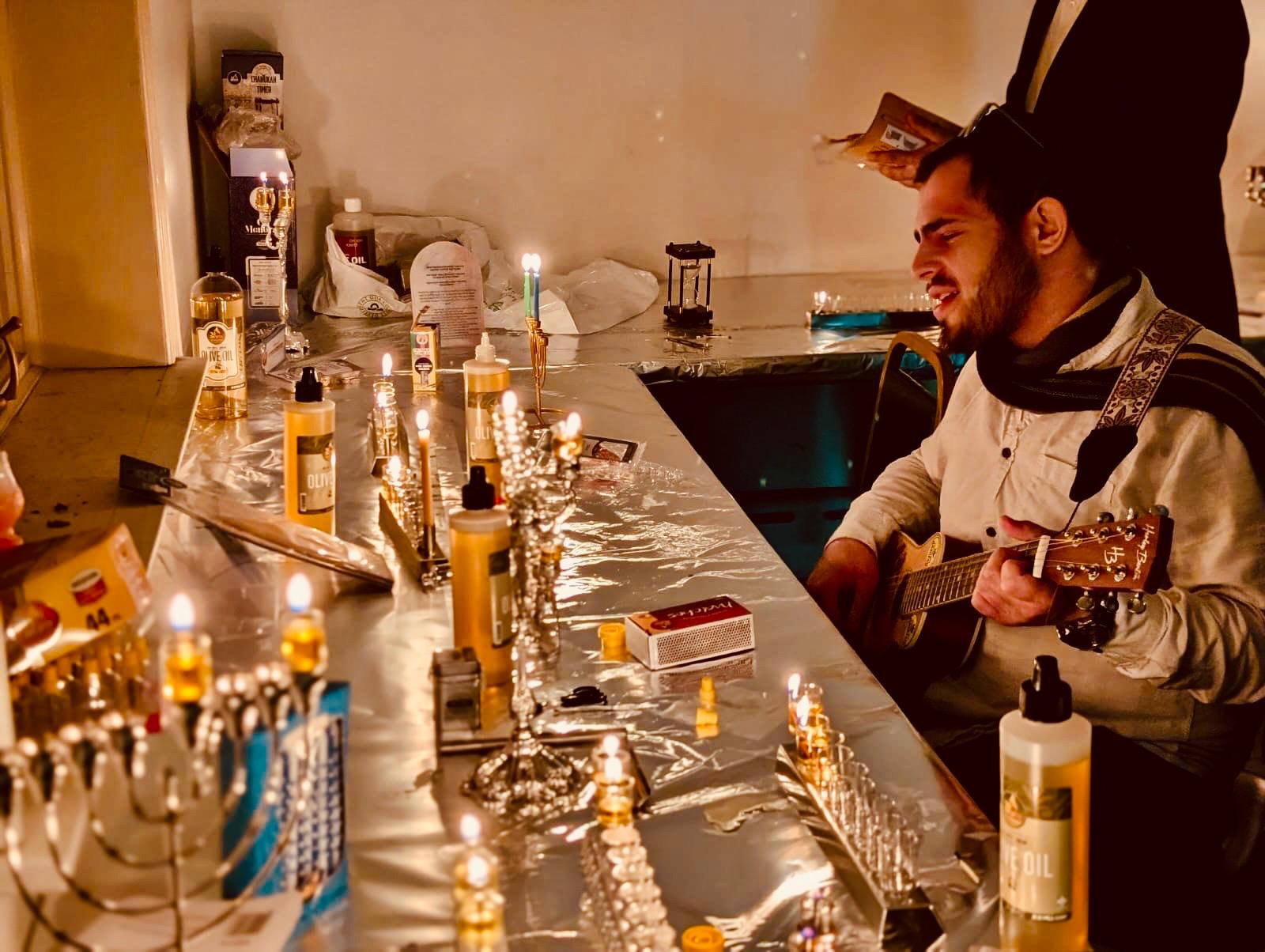 A talmid playing guitar beside a long row of lit menorahs