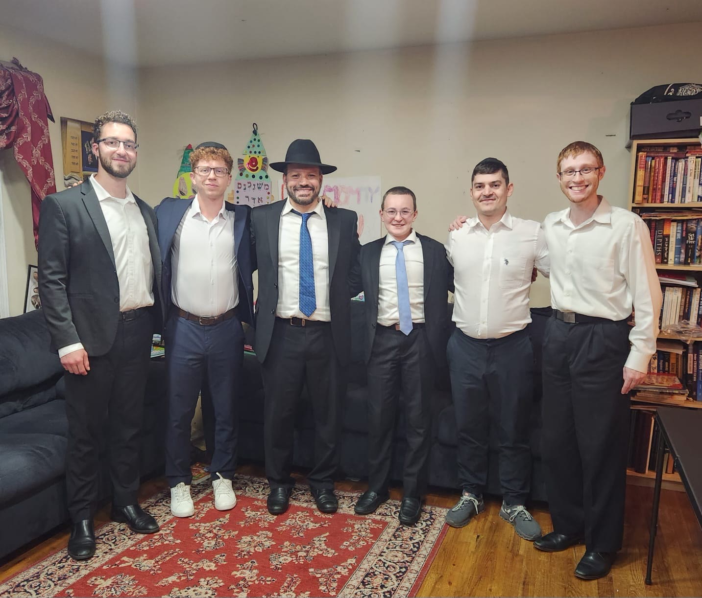 Six talmidim standing together with a rebbe in his living room