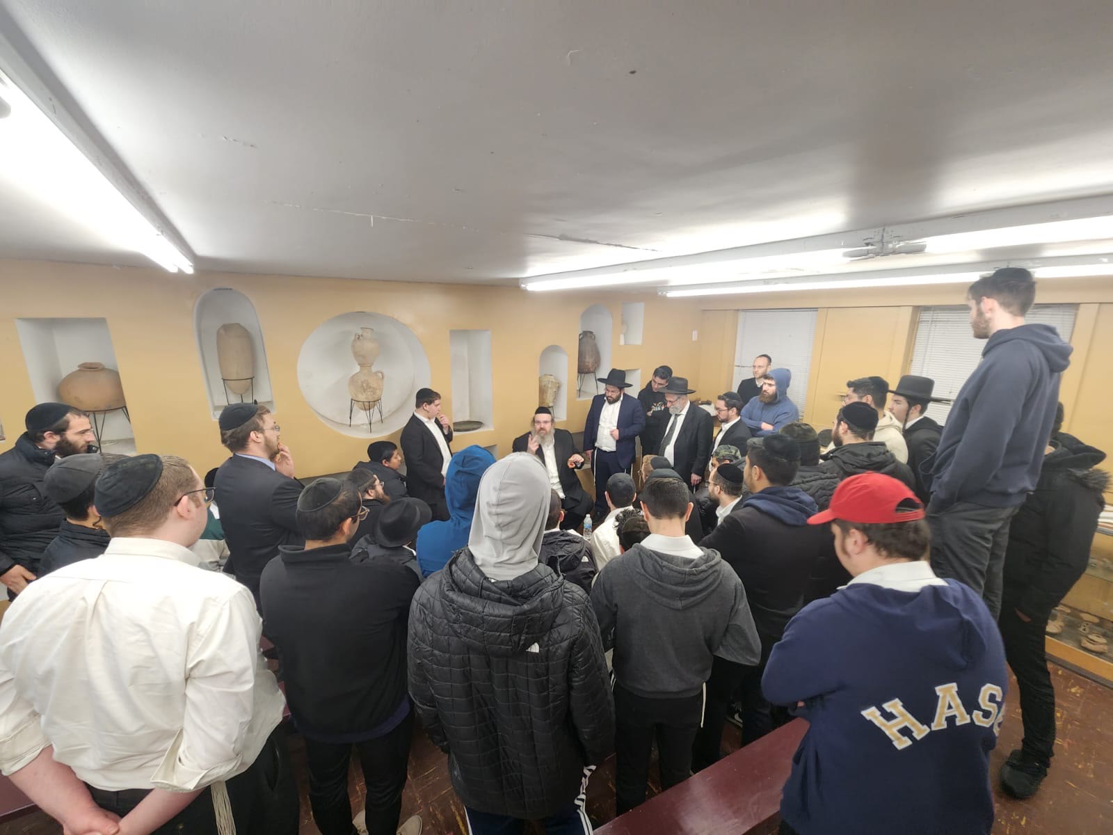 Talmidim gathered around a rebbe in a room displaying ancient clay vessels