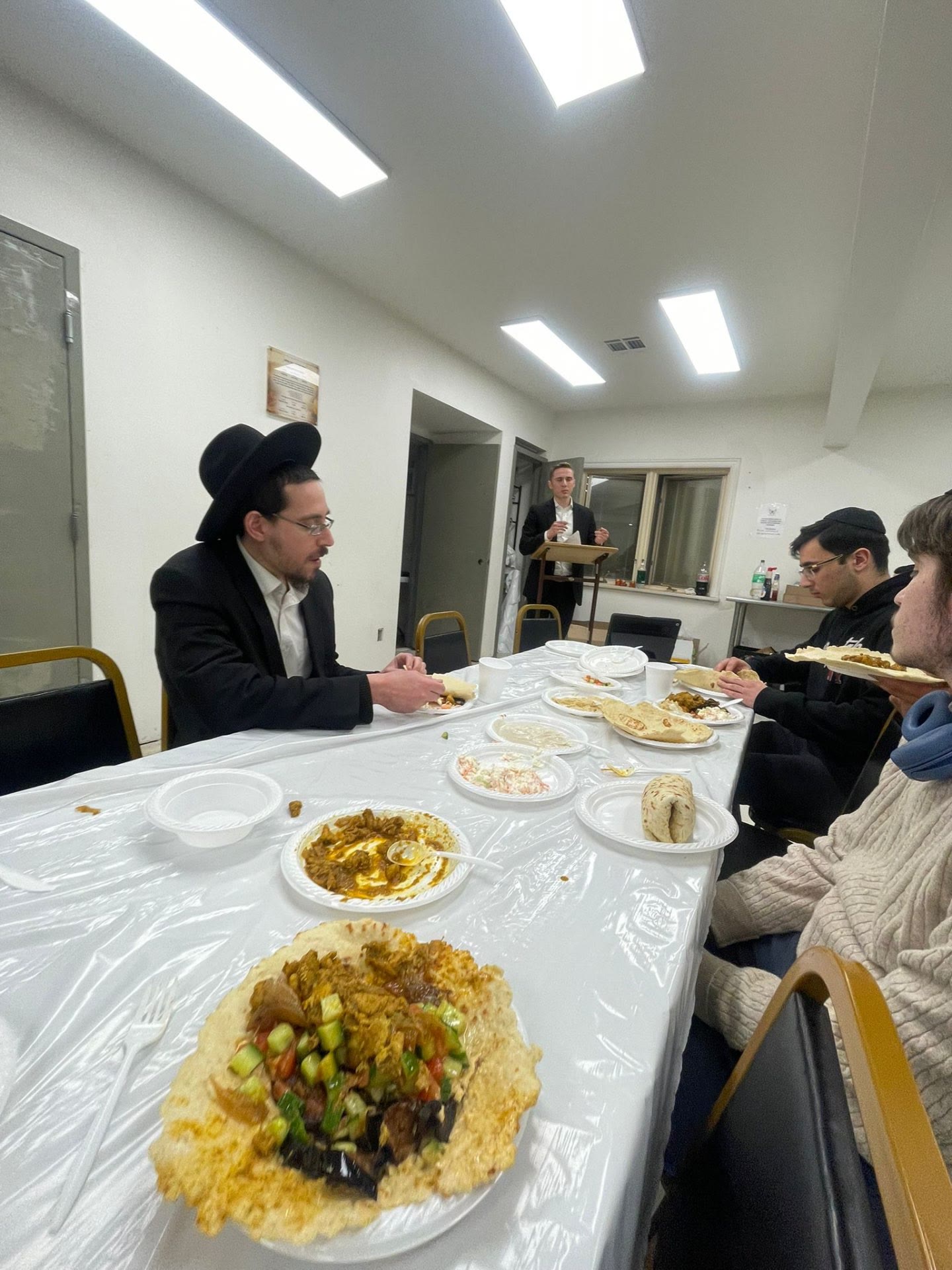 Talmidim eating together while a speaker addresses the room
